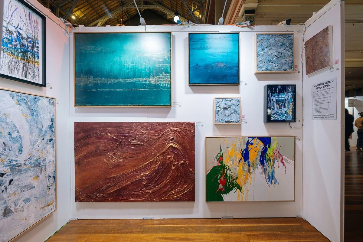 Collection of abstract paintings displayed on white gallery walls, featuring various textures, colors, and styles, with some artworks having prominent blues, reds, and metallic accents.