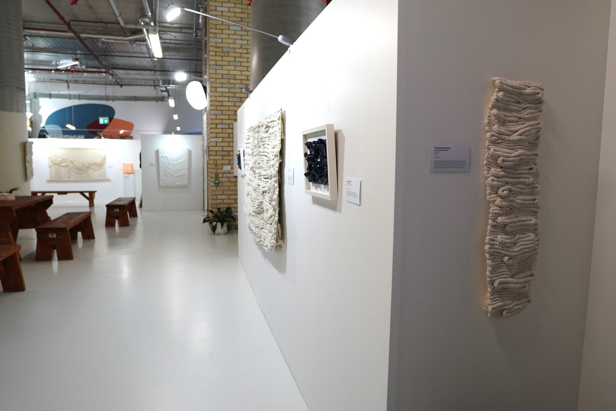 Contemporary art gallery with textured textile wall hangings and sculptures on white walls, wooden benches, and a brick wall in the background.