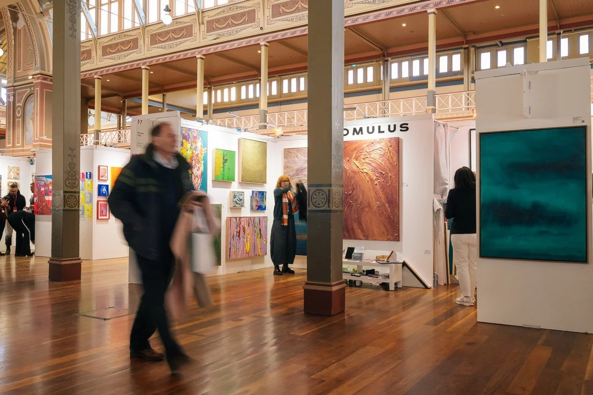 An art gallery with various colorful abstract paintings displayed on white walls, visitors observing the artwork, in a spacious room with high ceilings and wooden floors.