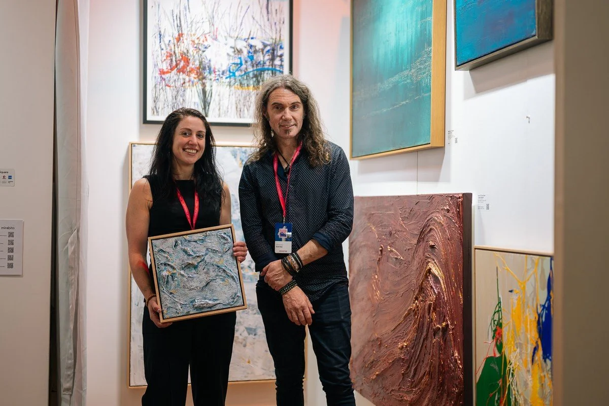 Woman and man standing in an art gallery, holding an abstract painting, surrounded by various colorful artworks on the walls.