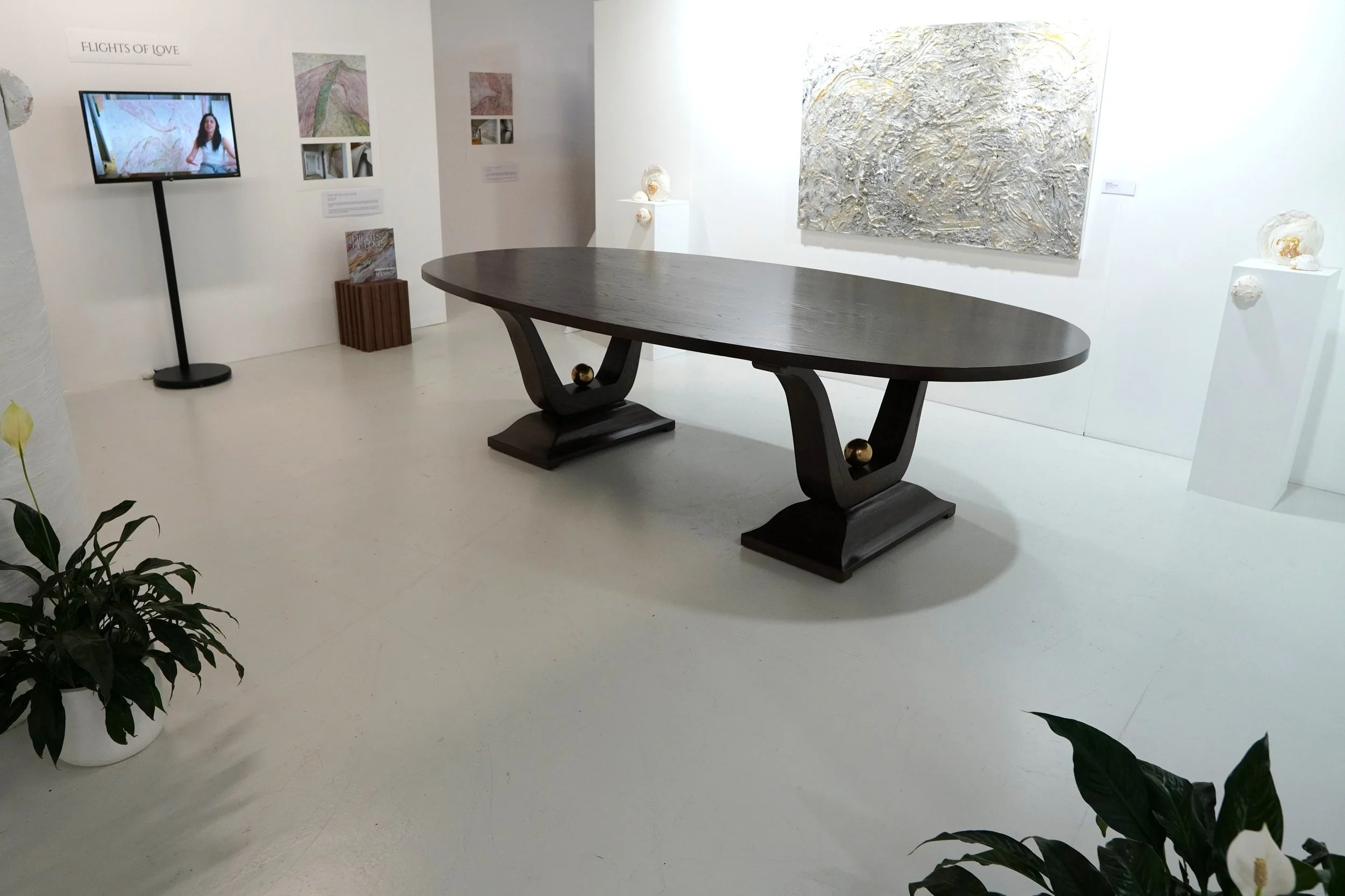 An art gallery with a large dark wooden table with gold-colored balls at the base of the legs, white walls with artwork, a small TV on a stand, and potted plants in the foreground.