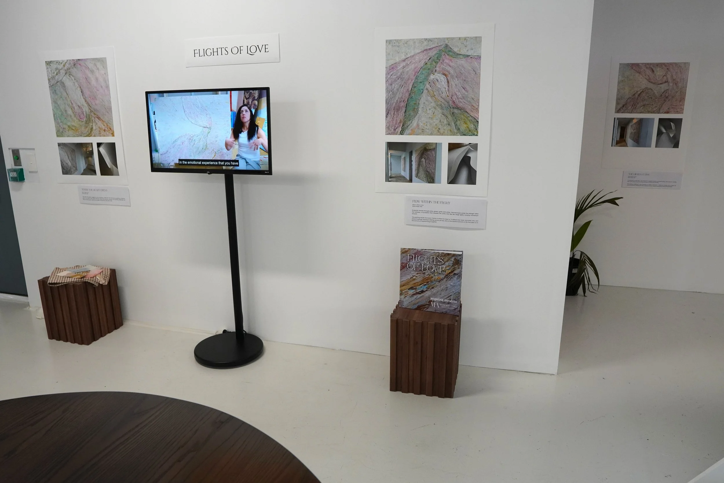 Gallery exhibit showcasing artwork titled 'Flights of Love' with informational plaques, a book display, and a woman explaining the artwork on a monitor.