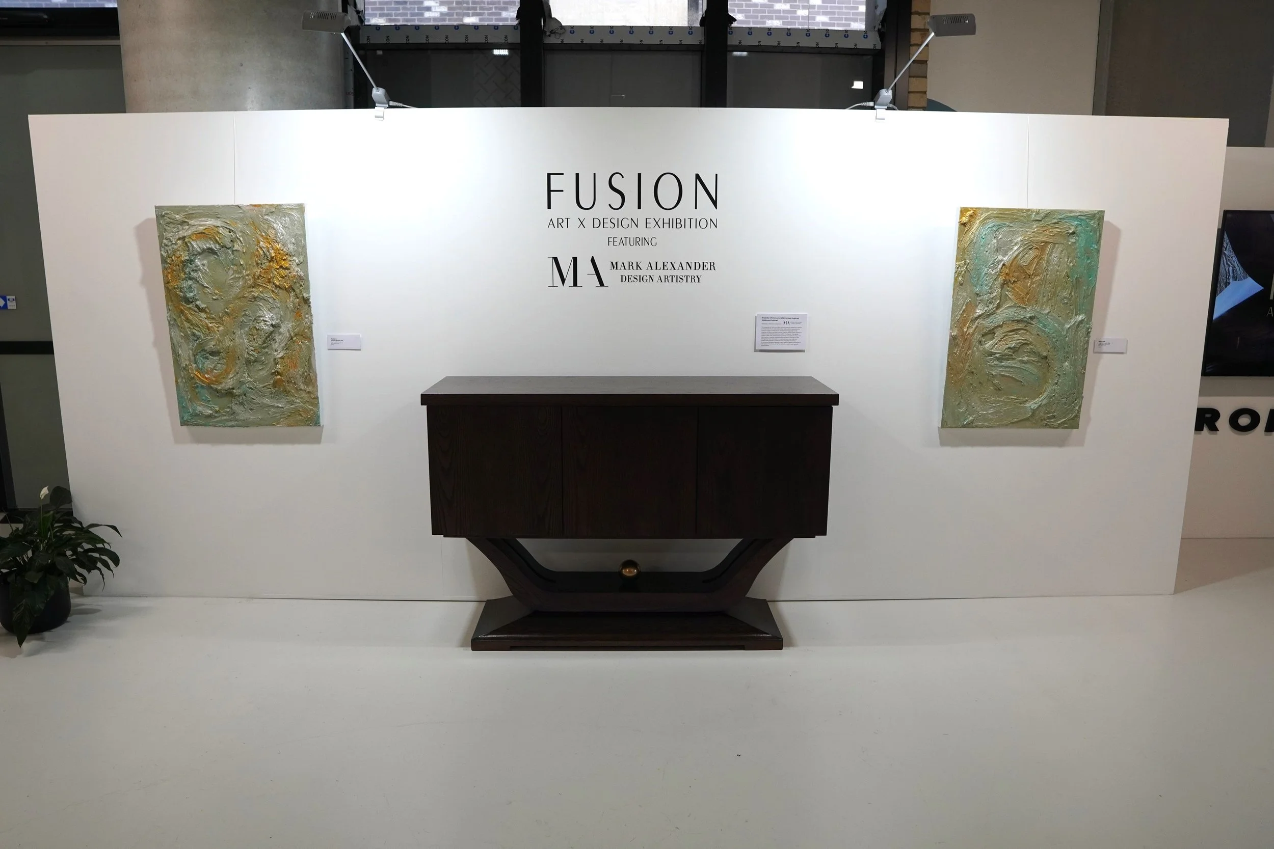 Exhibition display wall for the Fusion Art x Design Exhibition featuring two textured abstract paintings and a dark wooden side table in the center.