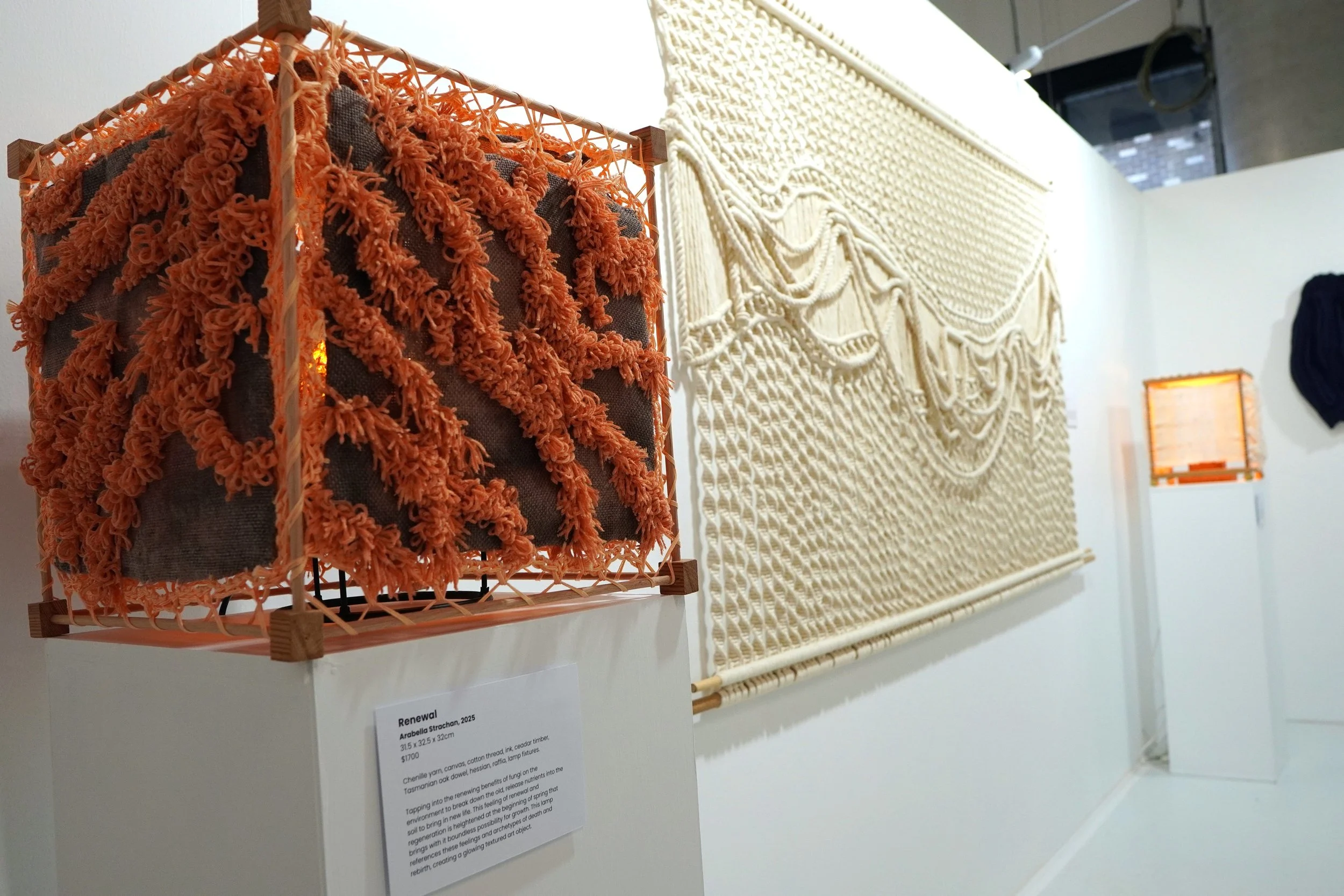 Two illuminated textile art pieces displayed on a white gallery wall. The piece on the left is a cube with orange, shaggy yarn pom-poms over a dark background, while the piece on the right is a large, intricate, cream-colored macramé wall hanging wit