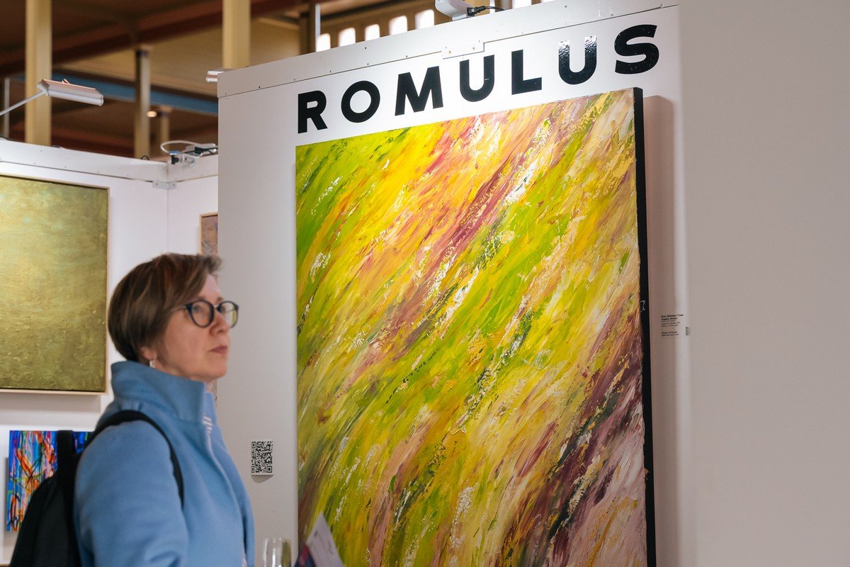 A woman with short hair, glasses, and a blue jacket looks at an abstract painting with yellow, green, pink, and brown brushstrokes at an art exhibit.