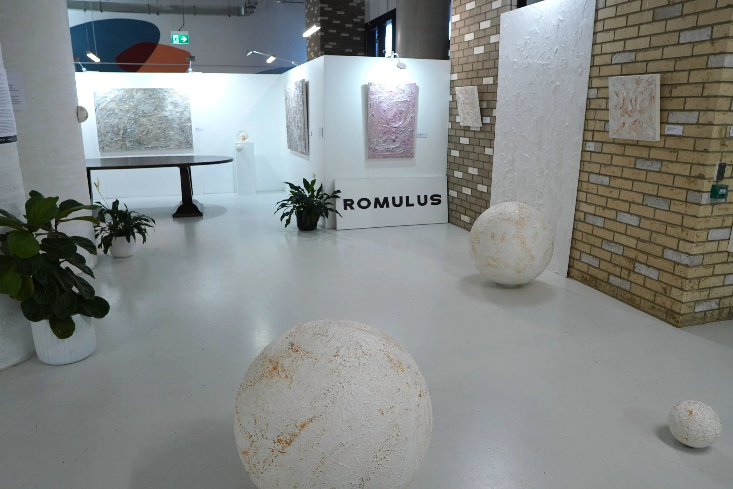 An art gallery display with abstract paintings on the walls, potted plants, and large decorative spheres on the floor. A sign reads 'ROMULUS' in bold black letters.