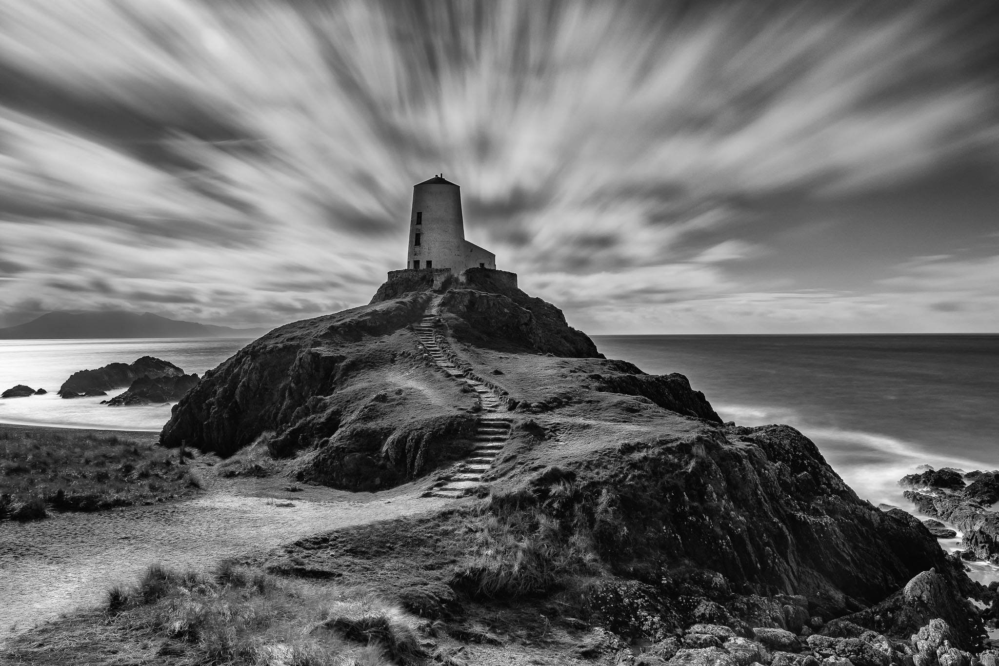 A monochrome image of a llanddwyn island lighthouse standing tall on a rugged cliff overlooking the sea. (Copy)