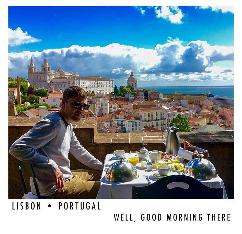 A man enjoying breakfast at an outdoor terrace overlooking Lisbon, Portugal, with a view of the city rooftops, the Jerónimos Monastery, and the ocean in the background.