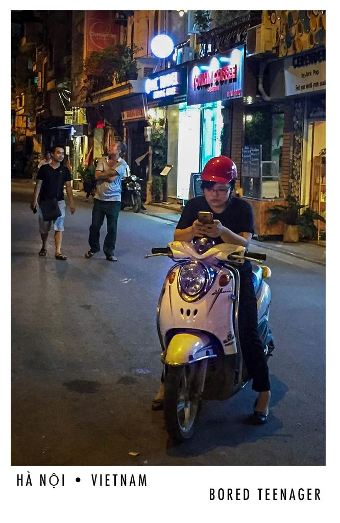 A young woman wearing a red helmet, sitting on a scooter and using her phone on a city street at night in Hanoi, Vietnam, with a few pedestrians and illuminated storefronts in the background.