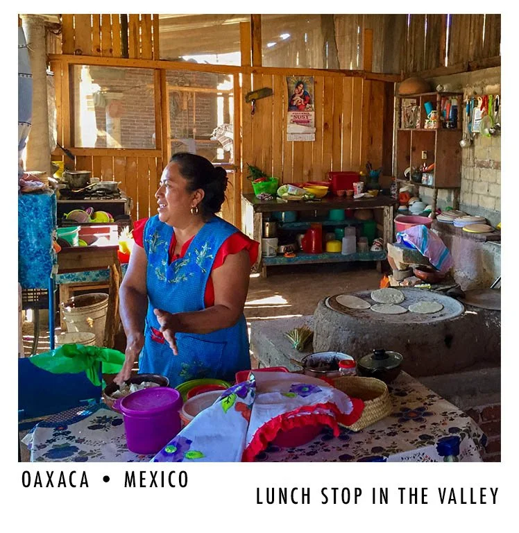 A woman in a blue and red traditional outfit standing in a rustic kitchen with colorful dishes and cooking utensils. She is smiling and appears to be preparing or serving food, with a wood-burning stove in the background. The setting is a simple, rur