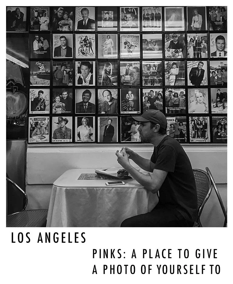 A man sits alone at a table in a restaurant with a wall of framed photographs behind him. The photo is in black and white. There is a caption that reads: "Los Angeles - Pinks: A place to give a photo of yourself to."
