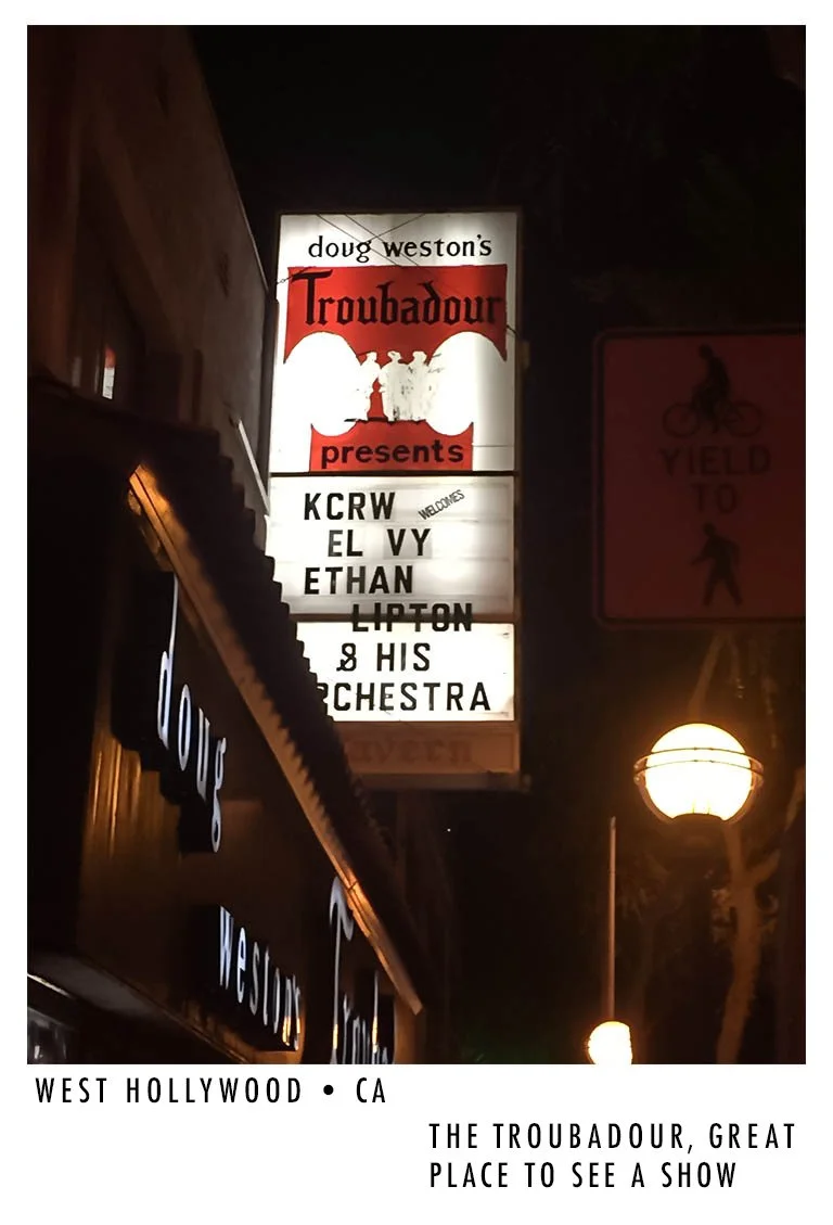 Illuminated marquee sign for Doug Weston's Troubadour music venue in West Hollywood, California, advertising a show with KCRW, El Vy, Ethan Lipton and his Orchestra, with some letters partially obscured.