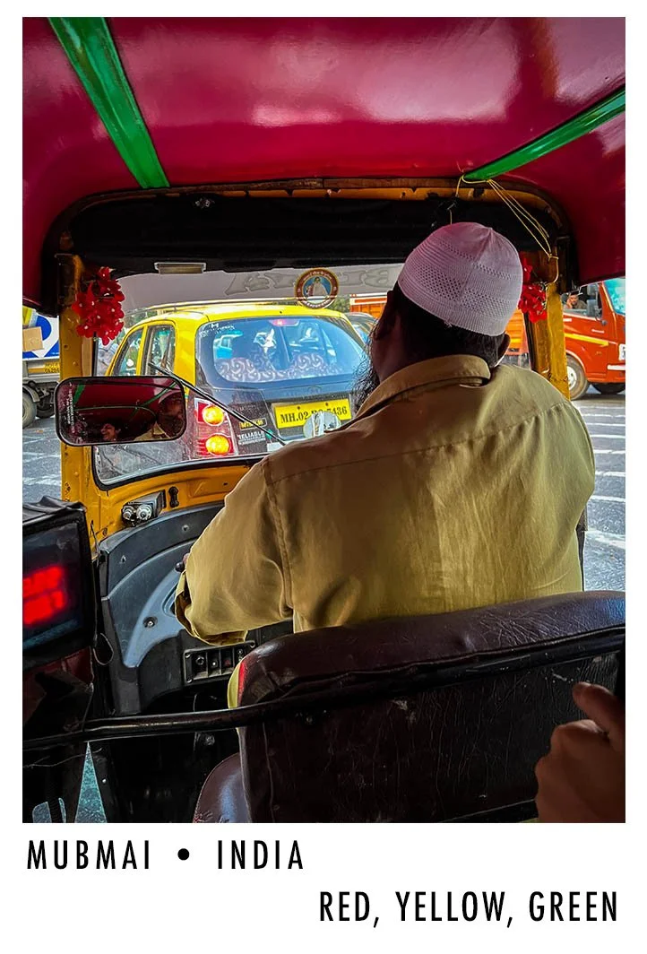 The photo shows the inside of an auto rickshaw in Mumbai, India, with a man wearing a white cap and yellow shirt driving. The view through the front includes cars on the road, and the dashboard is visible with some controls and a mirror.