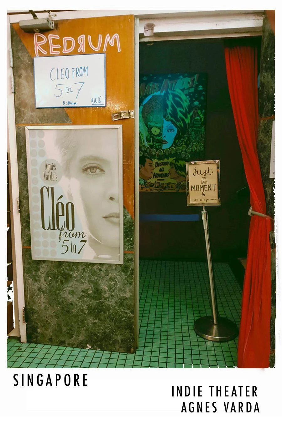 Entrance to Agnes Varda indie theater in Singapore with a poster for the film 'Cléo from 5 to 7' and a sign that says 'Just a Moment &'. Red curtain partially covers the entrance, and there is a colorful poster with a woman's face in the background.