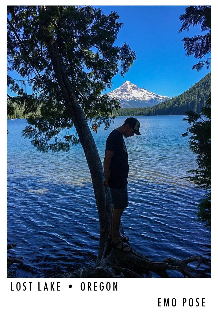 A person standing on a tree root near a lake in Oregon, with a snow-capped mountain in the background, and a clear blue sky.