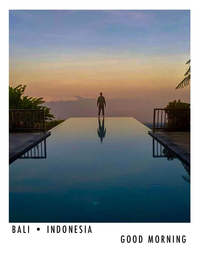 A person standing by the edge of an infinity pool at sunrise or sunset with a scenic view of the sky and landscape in Bali, Indonesia.