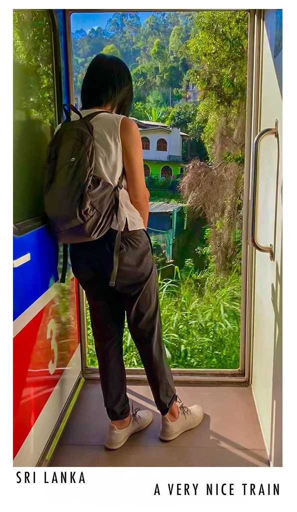 Young woman with black hair, wearing a sleeveless top, gray backpack, black pants, and gray sneakers, standing inside a train near the door, looking outside at lush green trees and a hillside with houses.