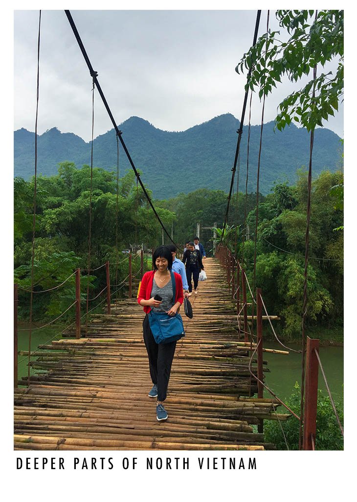 A woman walking on a rustic wooden suspension bridge surrounded by lush green trees with mountains in the background in North Vietnam.