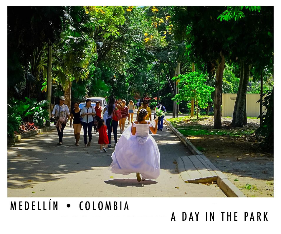A girl in a white dress appears to be running or dancing in a park with a paved walkway, surrounded by green trees, with a group of diverse people walking in the background.