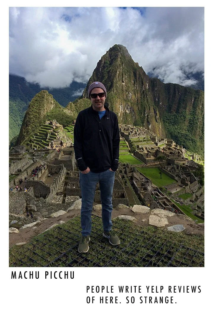 A man wearing sunglasses, a beanie, and a black jacket standing in front of Machu Picchu with mountains and clouds in the background.