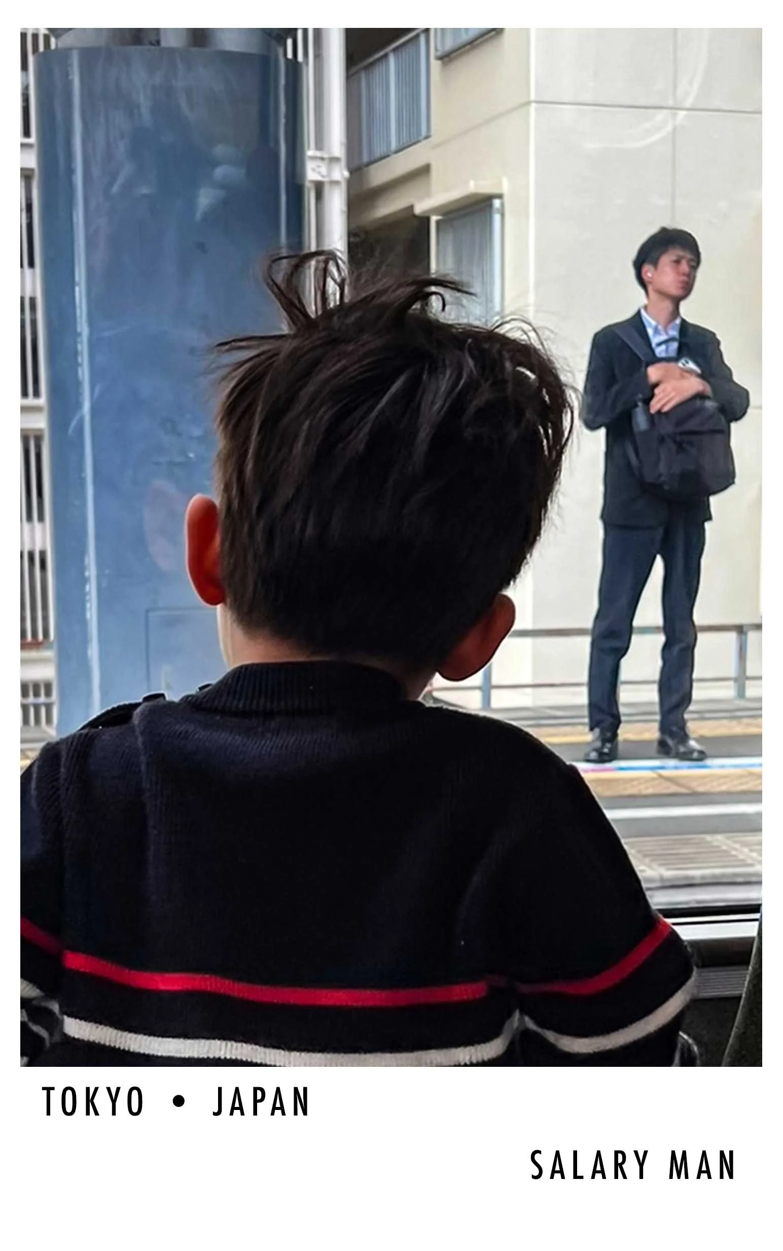 A young boy with dark hair view from behind, watching a man in a suit on a stage in a city environment. The boy wears a black jacket with red and white stripes, and the man on stage holds a backpack.