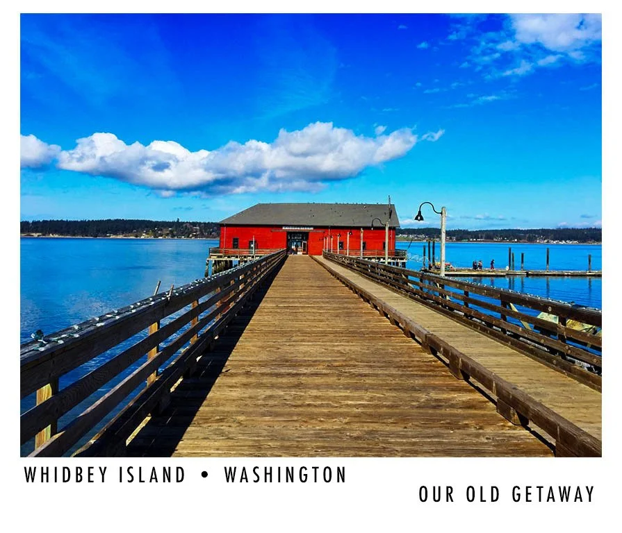 A wooden dock extends out over water toward a red building on stilts, with a cloudy blue sky and distant shoreline in the background, representing Whidbey Island in Washington.