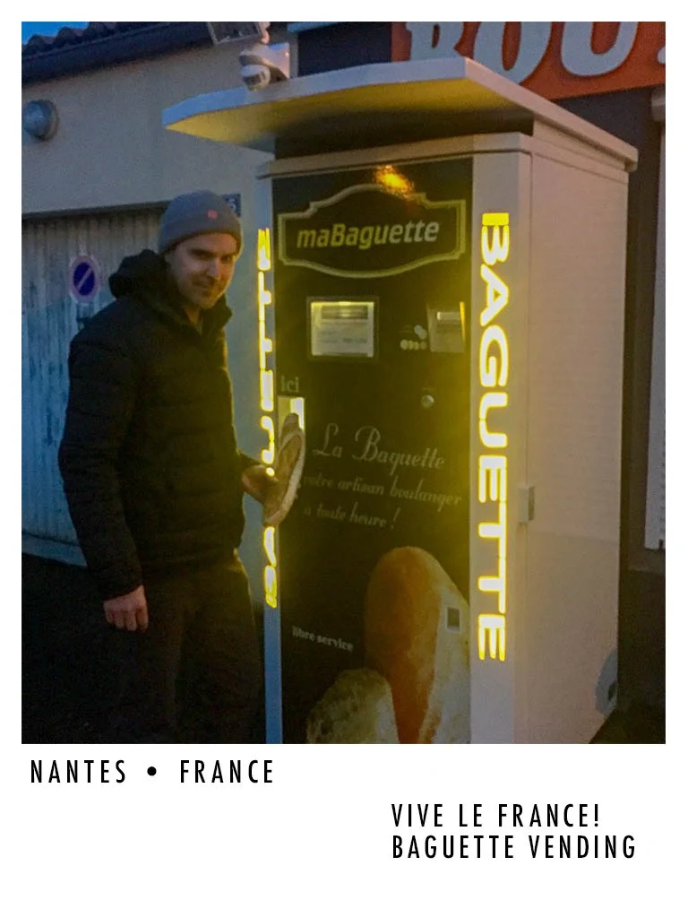 A man in a gray beanie and black jacket standing next to a vending machine for baguettes in Nantes, France, with illuminated yellow text and a baguette advertisement.
