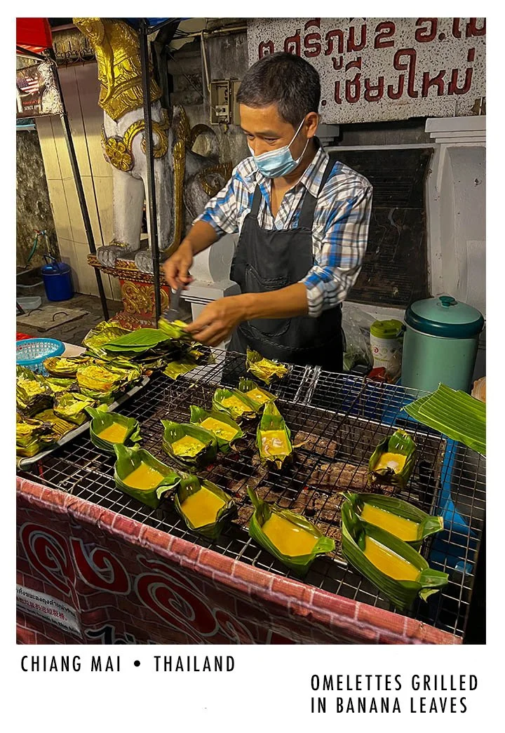 A man wearing a mask and apron preparing grilled omelettes wrapped in banana leaves at a street food stall in Chiang Mai, Thailand.