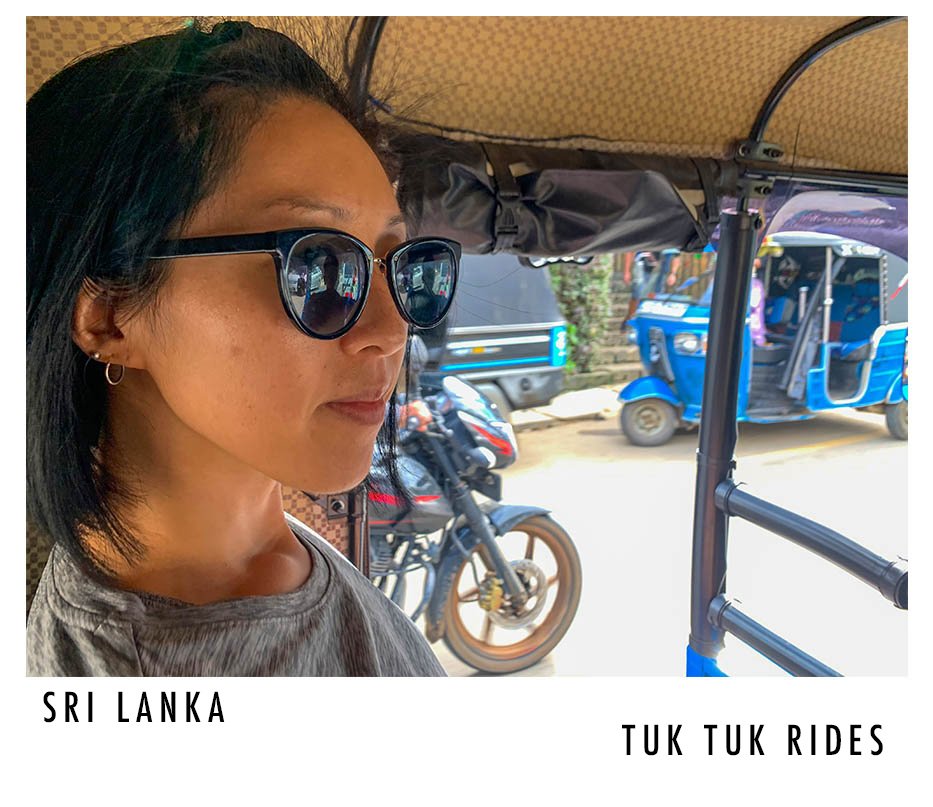A woman with black hair, wearing dark sunglasses and small hoop earrings, sitting inside a tuk tuk in Sri Lanka. The background shows a busy street with motorcycles, tuk tuks, and people.