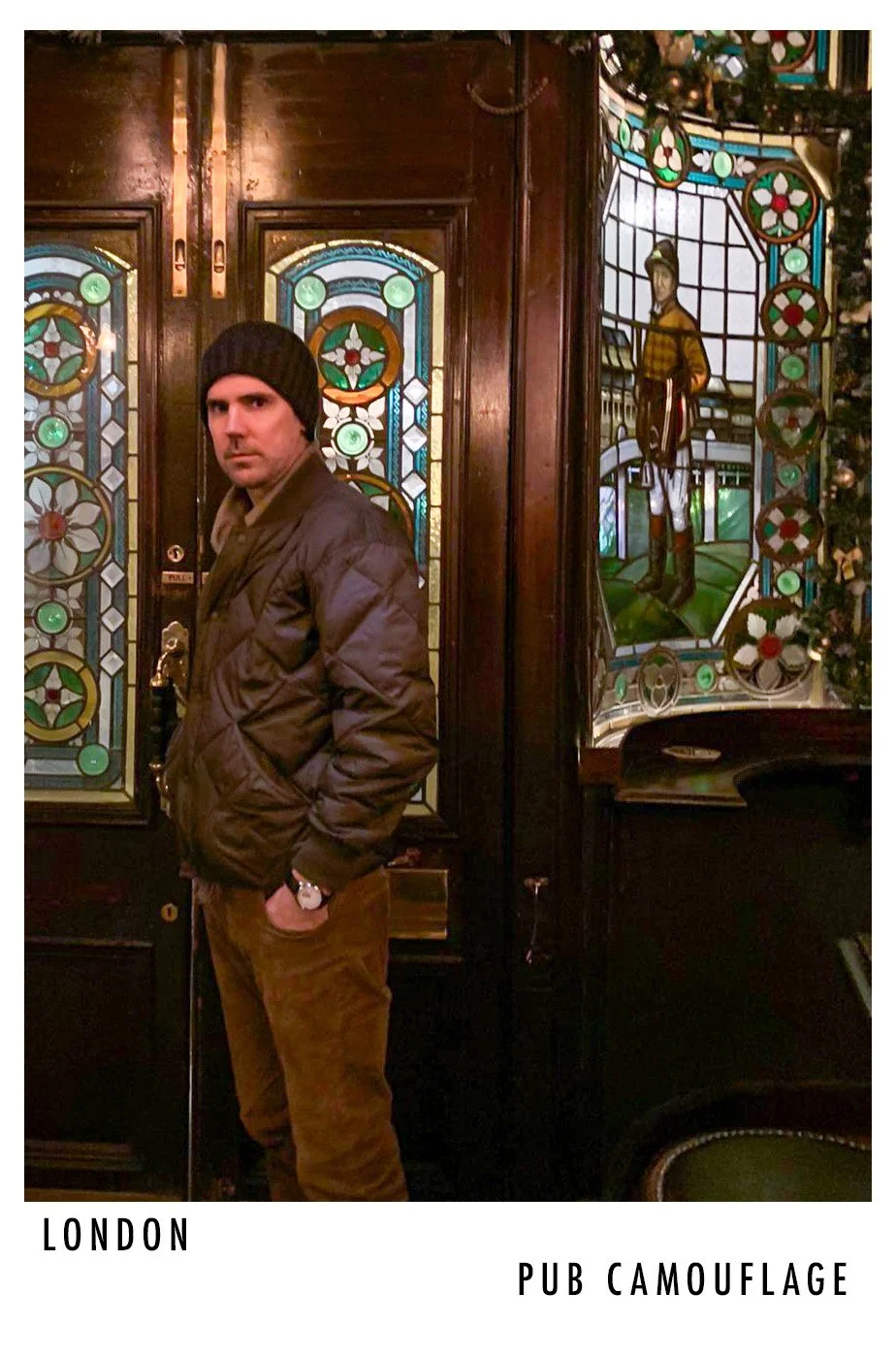 Man standing inside a pub in London, wearing a black beanie, brown pants, and a dark jacket, with stained glass windows and stained glass artwork in the background.