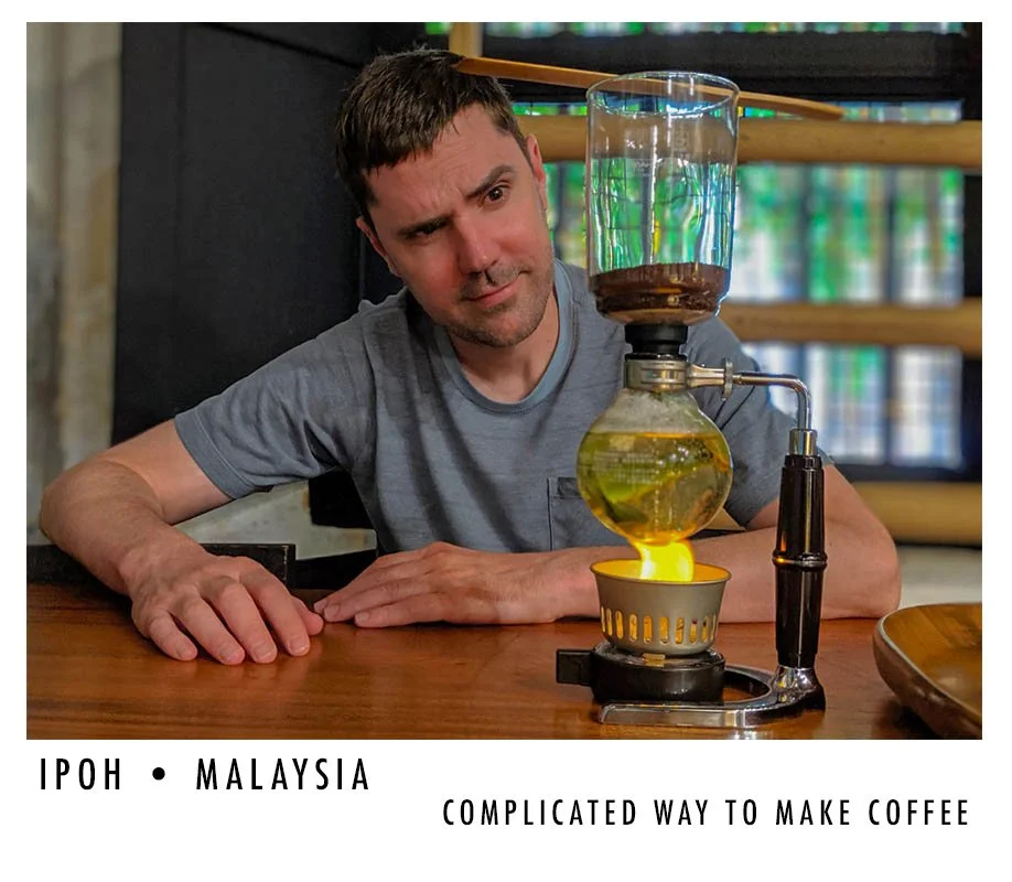 A man observing a complex coffee brewing process with multiple glass chambers and a flame underneath, on a wooden table in a coffee shop.