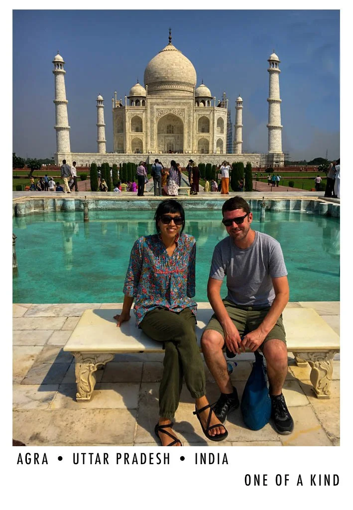 Tourists sitting on a bench in front of the Taj Mahal, Agra, Uttar Pradesh, India, with many visitors in the background and the monument's iconic white marble domes and minarets visible.