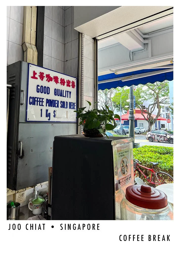 Interior of a coffee shop with a sign in both Chinese and English advertising coffee powder for sale. Outside view through the window shows trees, cars, and bicycles in Singapore.