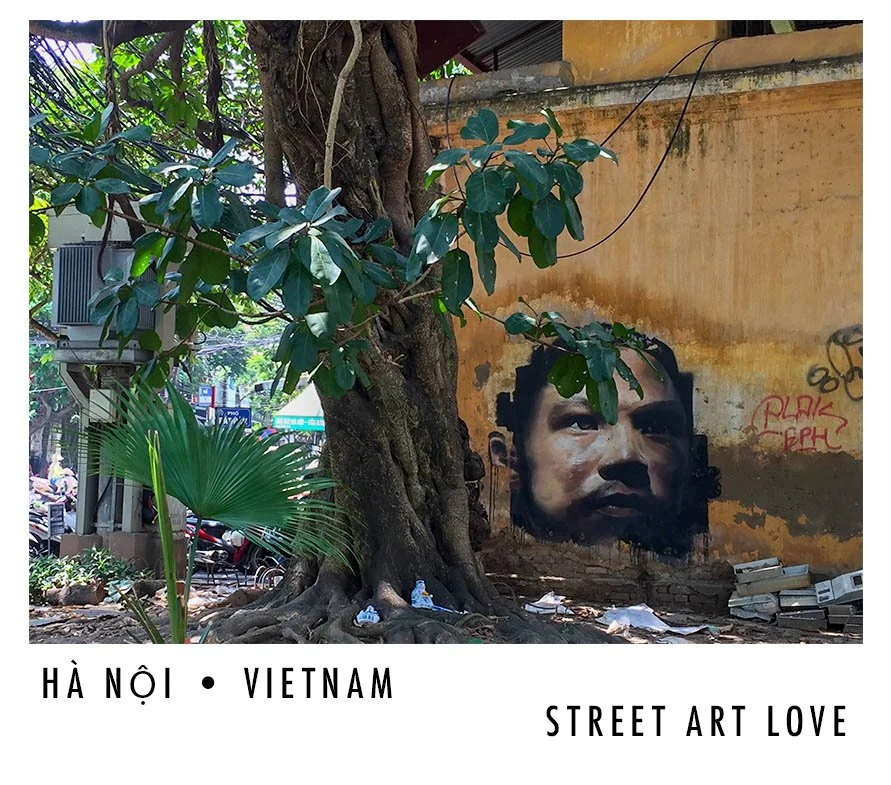 A mural of a child's face painted on a weathered yellow wall, partially covered by a large tree with lush green leaves in Hanoi, Vietnam.