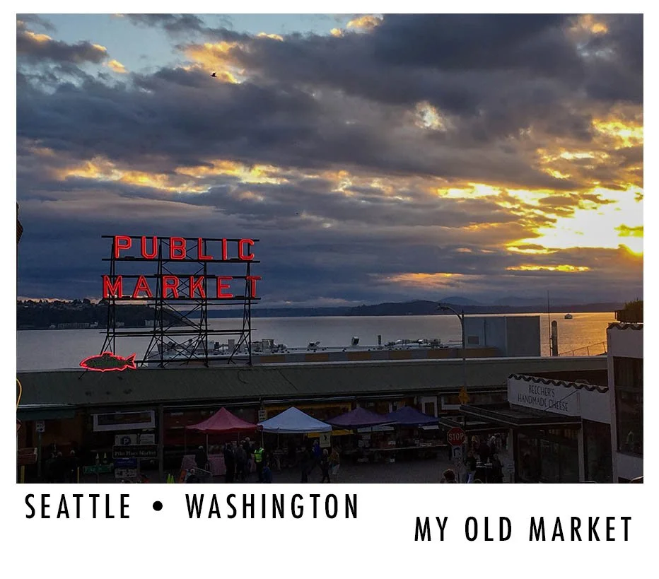Sunset over a waterfront public market in Seattle, Washington, with a neon sign reading 'Public Market' above the market. The sky is partly cloudy with the setting sun casting a warm glow, and people are gathered in front of the market.