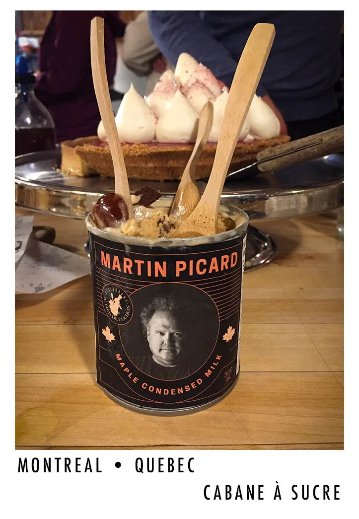 A tin of Martin Picard maple condensed milk ice cream with four wooden spoons inserted. In the background, there is a tart topped with whipped cream and some people standing behind. The text at the bottom reads "Montreal, Quebec, Cabane à sucre."
