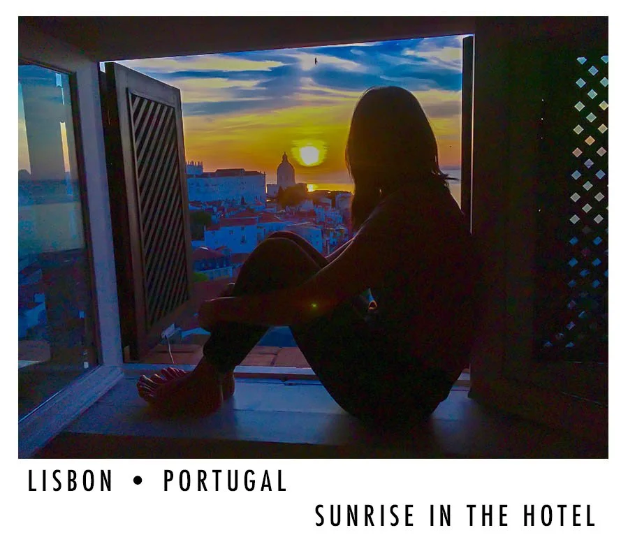 A person sitting by a large window watching the sunrise over Lisbon, Portugal, from inside a hotel room.