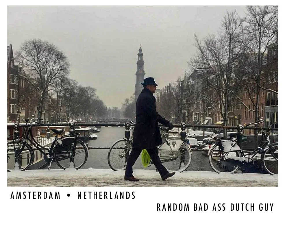 A man in dark clothing and a black hat walking along a snowy canal bridge in Amsterdam, Netherlands, with bicycles on either side and a church tower in the background, during winter.