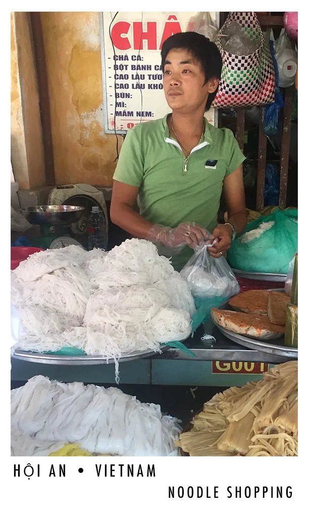 A woman at a noodle stall in Hanoi, Vietnam, with piles of white noodles and some cooked dishes on display.