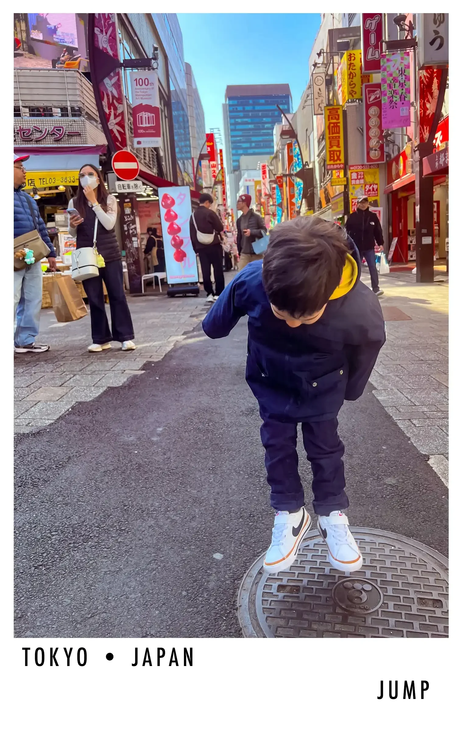 A young boy wearing white sneakers and black pants jumps onto a manhole cover on a busy Tokyo street with colorful signs and pedestrians in the background.