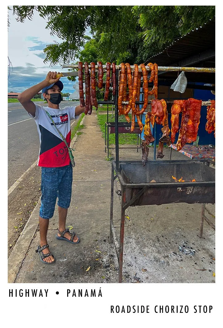 A boy wearing a black face mask, a cap, and casual clothes is grilling roadside chorizo sausages and pork ribs on a street in Panama, with a grill over open flames and sausage sticks hanging.