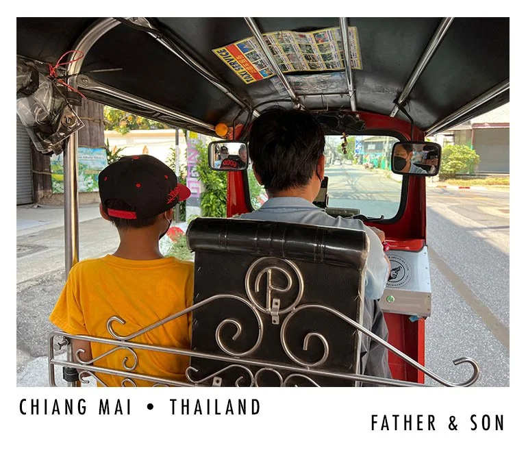 A man and boy riding in a tuk-tuk vehicle in Chiang Mai, Thailand, viewed from behind.