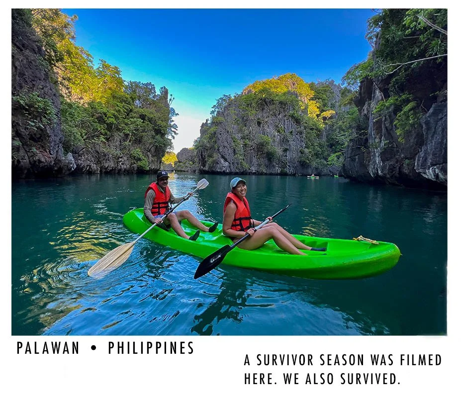 Two people kayak in a narrow, rocky canyon with lush greenery and clear blue water in Palawan, Philippines. Both are wearing red life jackets and smiling.