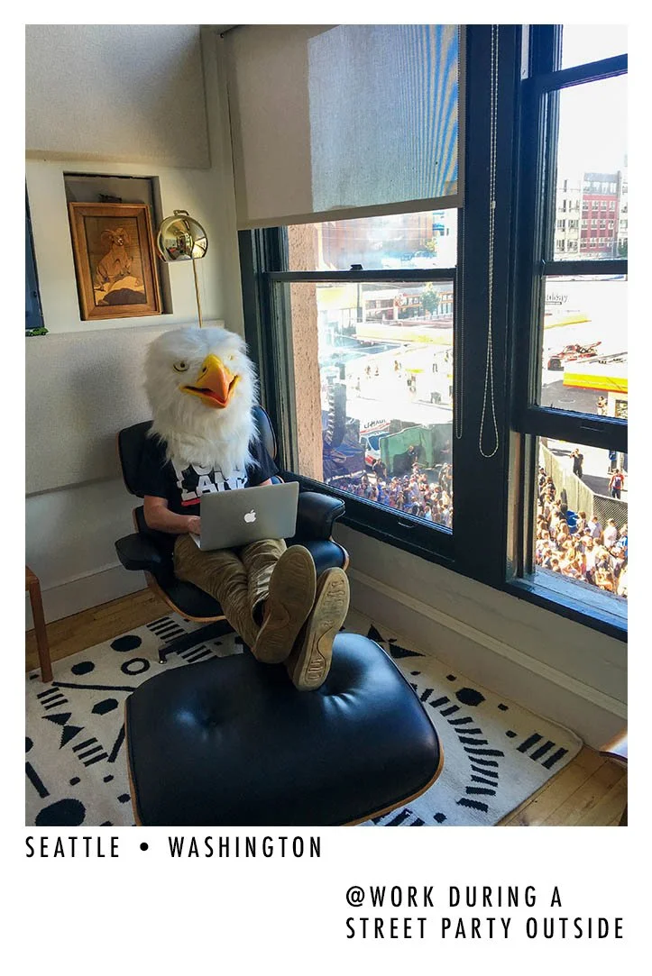 An indoor scene showing a person with a large eagle mask using a laptop while sitting in a modern black chair with matching ottoman next to a large window. Outside, a crowd is gathered on the street for a street party in Seattle, Washington.