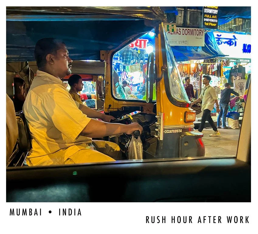 A man in a yellow shirt and dark trousers driving an auto rickshaw through a busy street in Mumbai, India during rush hour.