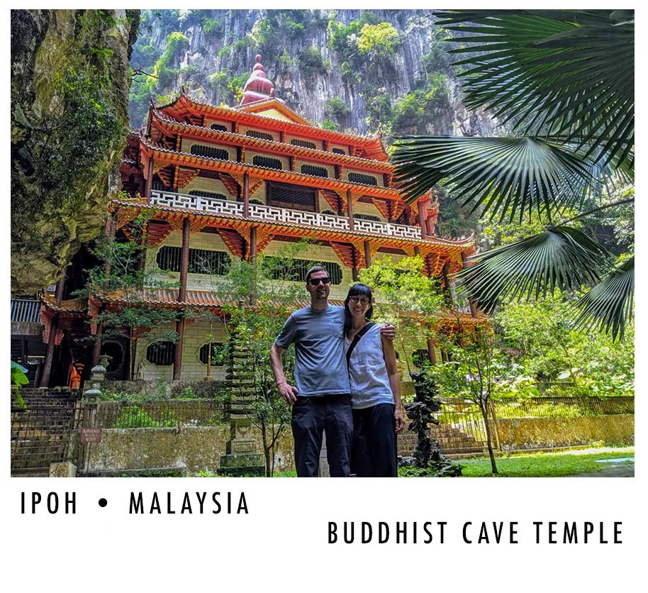 A couple standing in front of a large, ornate Buddhist cave temple building with multiple tiers and traditional Asian architectural features, surrounded by lush greenery and tall cliffs in Ipoh, Malaysia.
