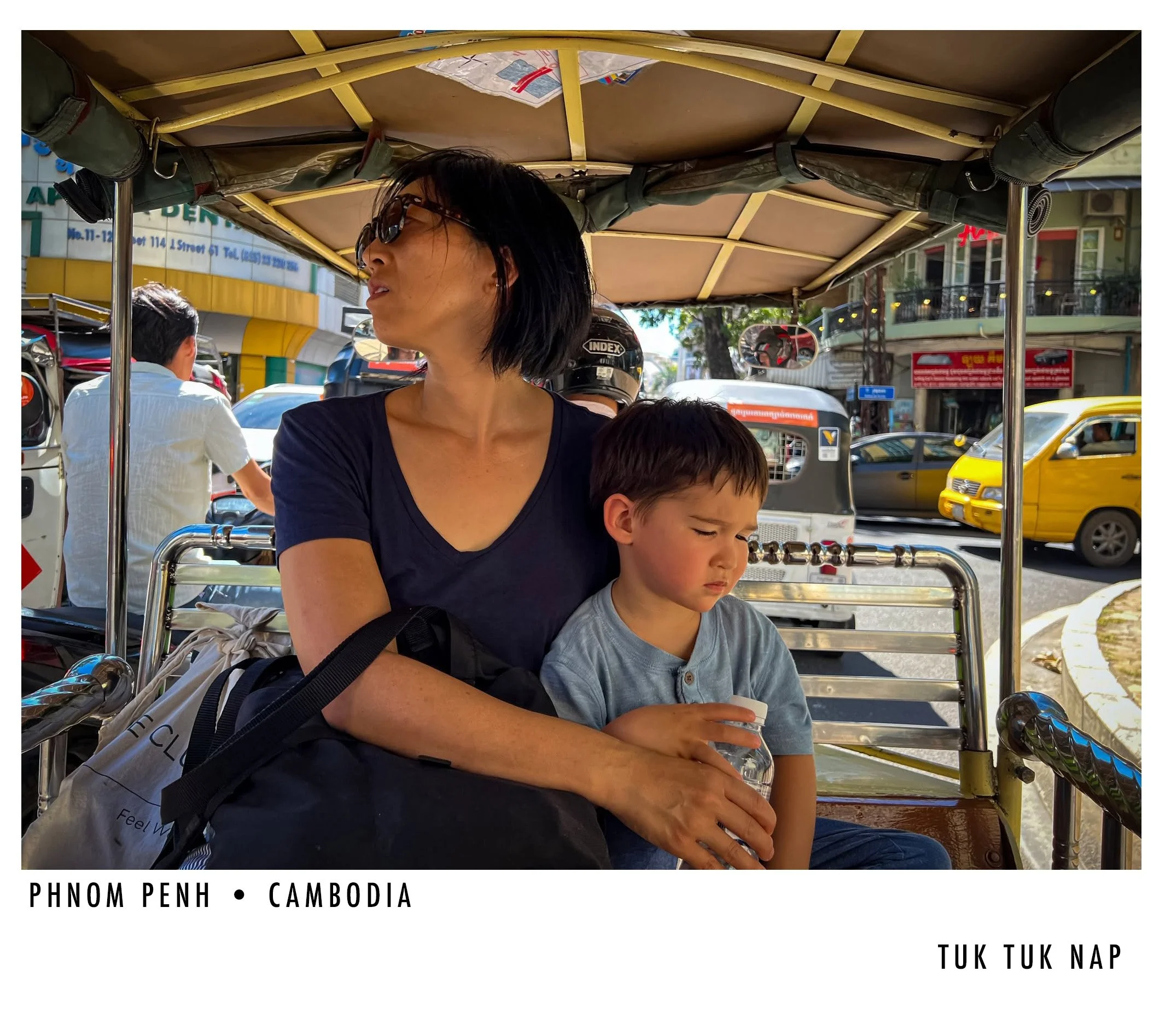A woman with black hair and sunglasses, sitting on a tuk-tuk in Phnom Penh, Cambodia, with a young boy beside her. The street is bustling with cars, motorcycles, and buildings in the background.
