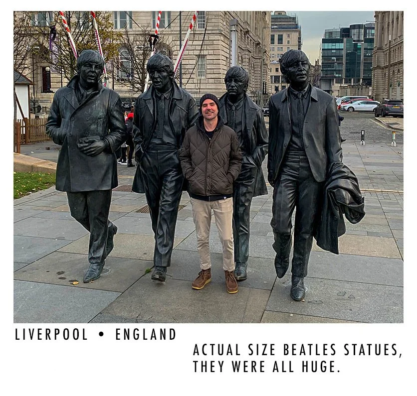 A man standing in front of the Beatles statues in Liverpool, England, surrounded by large statues of the Beatles members.