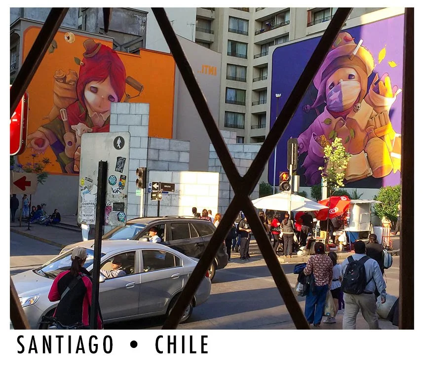 A busy street scene in Santiago, Chile, viewed through a decorative metal fence. The image features cars, pedestrians, and street vendors. Two large colorful murals of cartoon-like characters with large eyes and vibrant clothing are painted on buildi