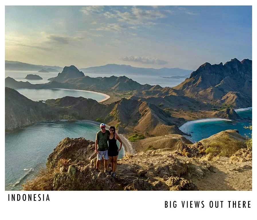 Two people standing on a rocky hilltop overlooking a scenic landscape of mountains, lakes, and ocean near Indonesia, with the caption 'BIG VIEWS OUT THERE'.